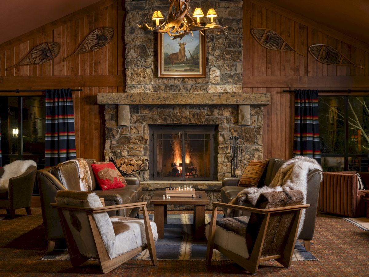 A cozy rustic living room with a stone fireplace, wooden walls, armchairs, fur throws, a chessboard, and vintage decor, including wall-mounted snowshoes.