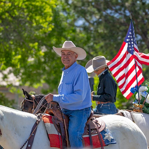 A smiling cowboy in a blue shirt rides a white horse, with another rider and an American flag in the background, sunny outdoor scene.
