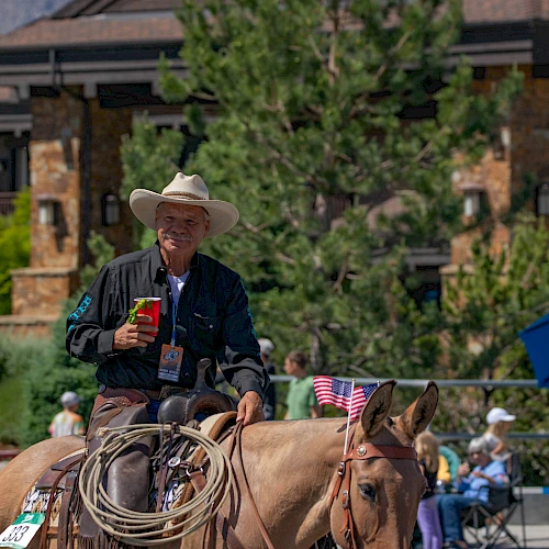 A man wearing a hat rides a horse with a lasso, holding a red cup, in a parade-like scene with onlookers and flags.