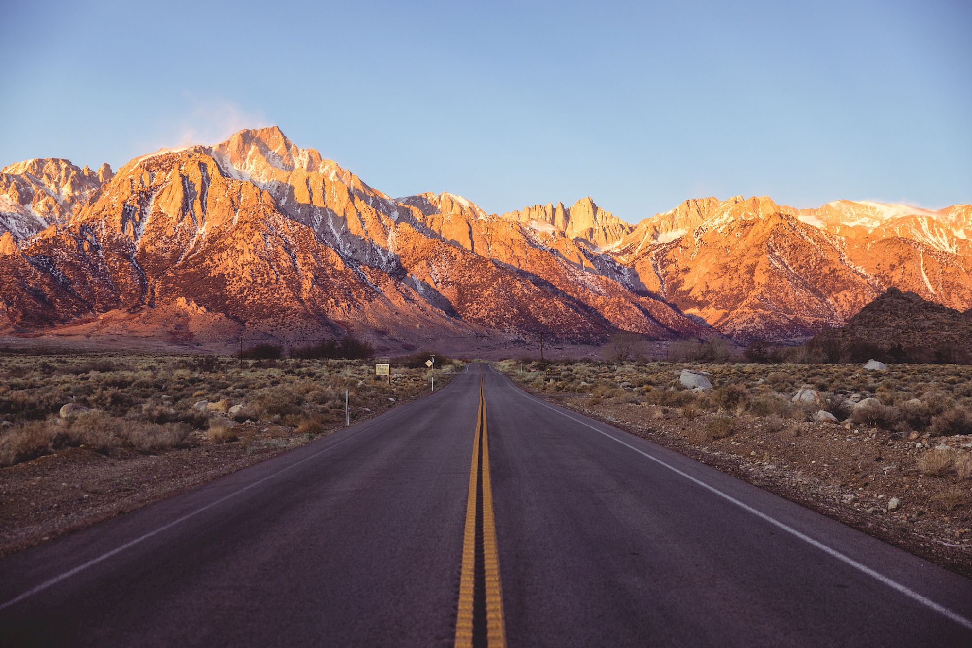 A long straight road leads toward rugged mountain peaks glowing orange in sunset light, desert scrub on either side, clear blue sky.