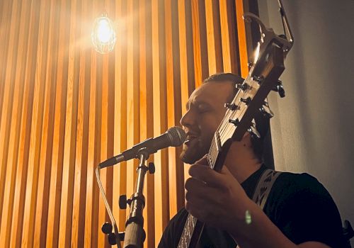 A person is playing a guitar and singing into a microphone. The background features a wooden panel with lighting.