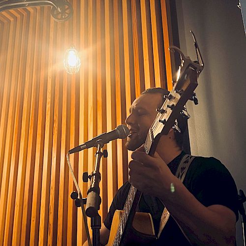 A person is playing a guitar and singing into a microphone. The background features a wooden panel with lighting.