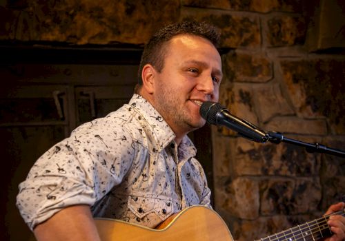 A person is playing an acoustic guitar and singing into a microphone in a cozy indoor setting.