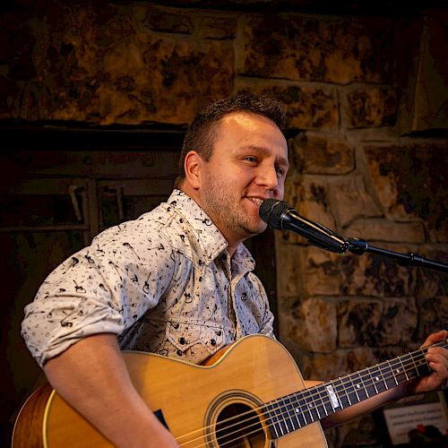 A person is playing an acoustic guitar and singing into a microphone in a cozy indoor setting.