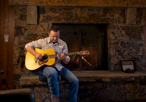 A person is sitting and playing a guitar in front of a large stone fireplace with a painting hung above it.