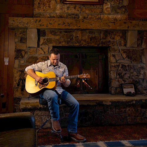 A person is sitting and playing a guitar in front of a large stone fireplace with a painting hung above it.
