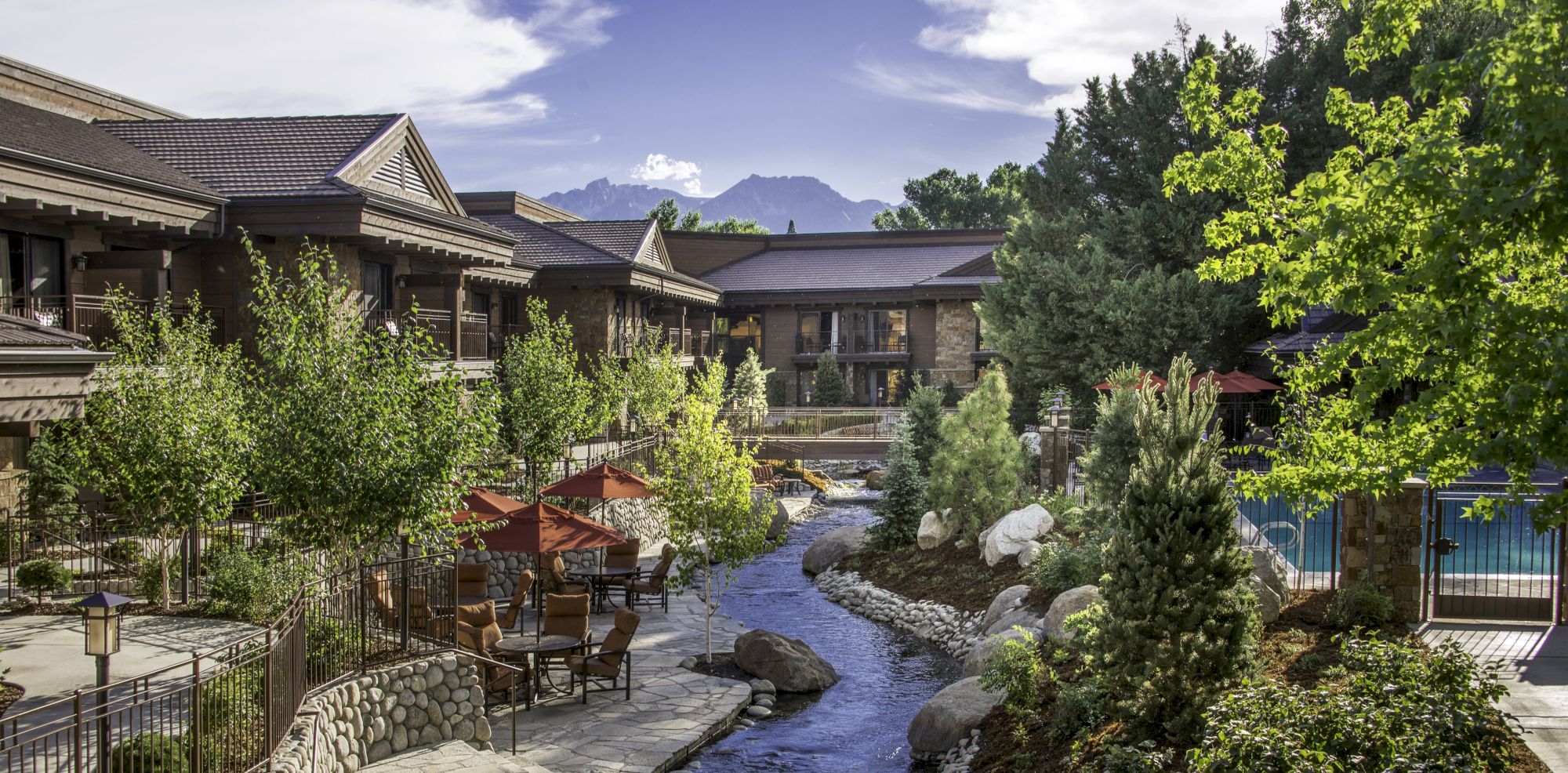 The image depicts a serene outdoor area featuring a creek surrounded by lush greenery, seating, and buildings with mountain views in the background.