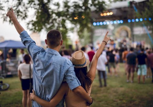 A couple, with arms around each other, enjoys an outdoor concert or festival, pointing towards the stage, surrounded by other attendees.