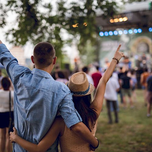 A couple, with arms around each other, enjoys an outdoor concert or festival, pointing towards the stage, surrounded by other attendees.