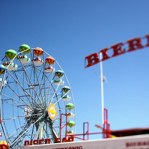 The image shows a vibrant Ferris wheel with colorful cabins, set against a clear blue sky. A "Bierk&ouml;nig" sign is visible in the foreground.