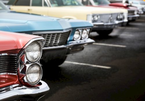 The image shows a close-up of the front ends of several classic cars parked in a row, with focus on their distinctive grilles and headlights.