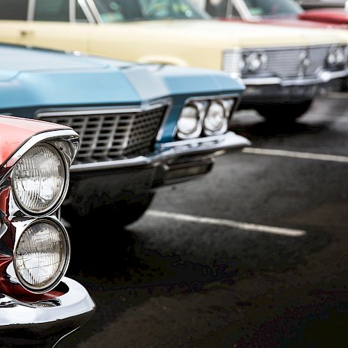 The image shows a close-up of the front ends of several classic cars parked in a row, with focus on their distinctive grilles and headlights.