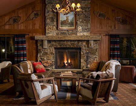 Cozy cabin living room with stone fireplace, antler chandelier, plaid curtains, armchairs, and a small table in front of a warm fire.