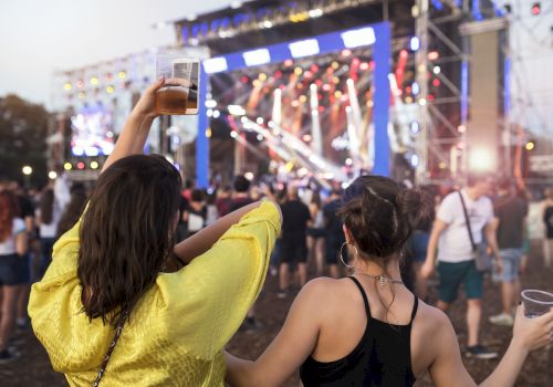 Two people enjoying a live outdoor concert, one raising a drink, with a brightly lit stage and a crowd in front of them.