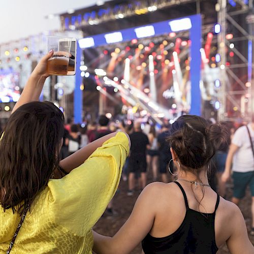 Two people enjoying a live outdoor concert, one raising a drink, with a brightly lit stage and a crowd in front of them.