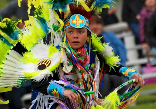 A person in vibrant traditional attire is performing a dance outside, with spectators in the background.