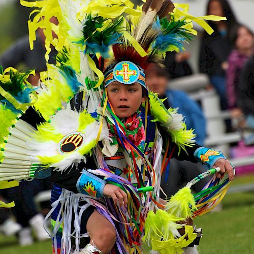 A person in vibrant traditional attire is performing a dance outside, with spectators in the background.