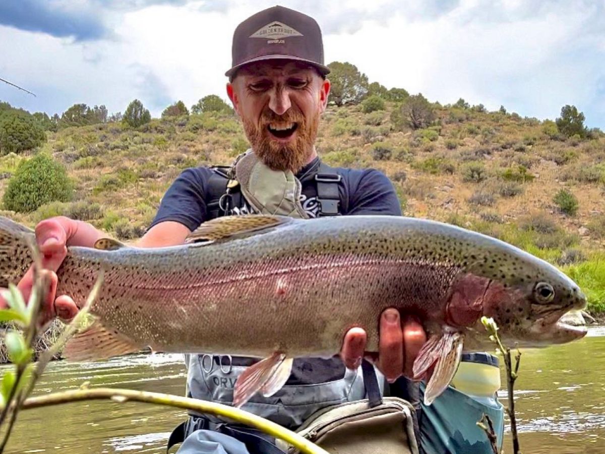 A man proudly holds a large fish near a river with a scenic, hilly background, appearing to smile or laugh.