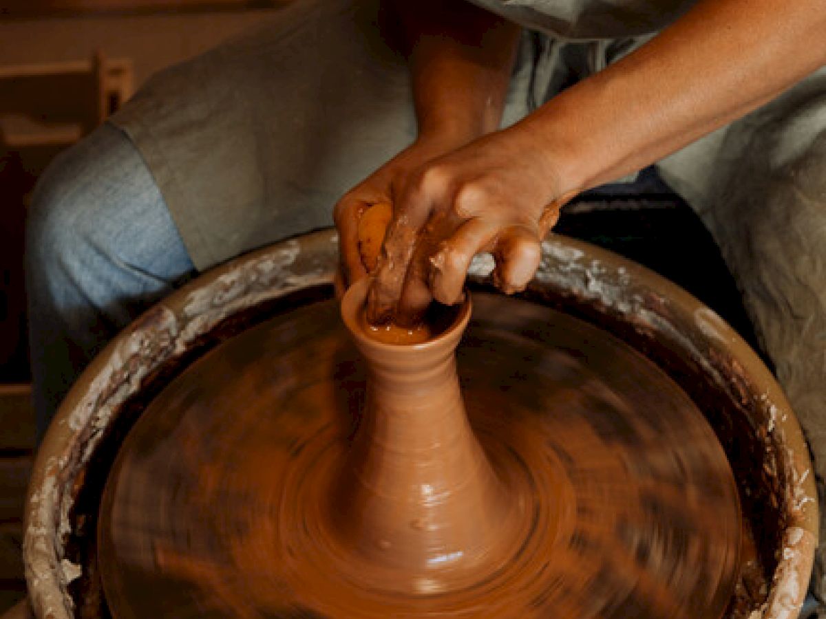 A person is using a pottery wheel to shape clay into a vessel. Hands are covered in wet clay while the wheel spins the pottery.