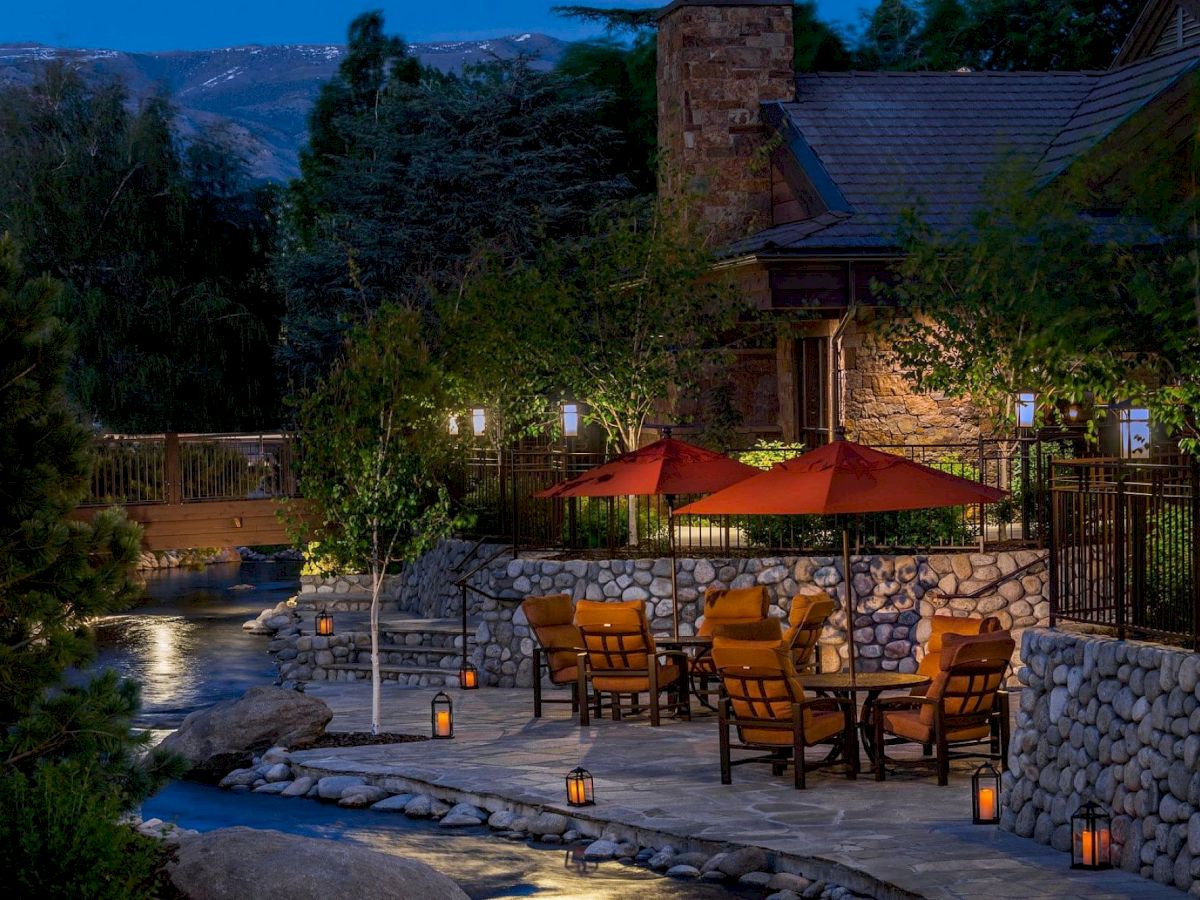 An outdoor patio with red umbrellas and seating next to a river, surrounded by trees and lanterns, near a stone building at dusk.