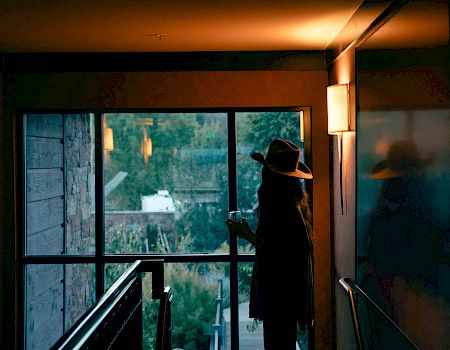 A person in a dark hat and coat stands indoors by a large window, lit by warm lamps, looking out at trees and dusk.