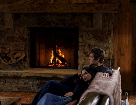 A man sits by a stone fireplace watching flames as a small child rests near him on a cozy blanket.