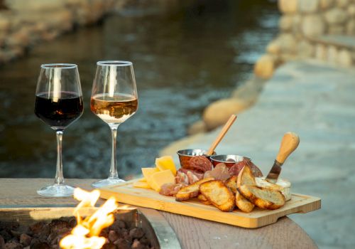 Two glasses of wine sit beside a cheese and meat platter near a fire pit, overlooking a serene stream with stones.