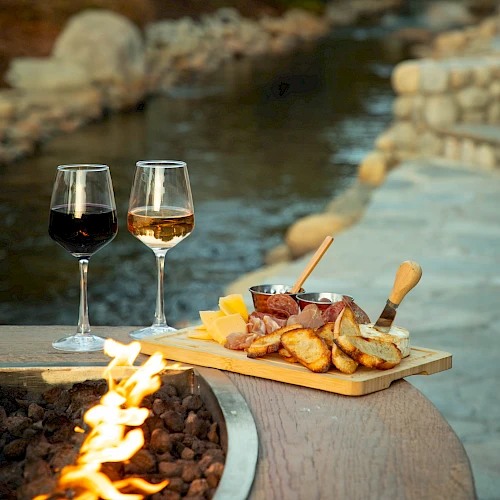Two glasses of wine sit beside a cheese and meat platter near a fire pit, overlooking a serene stream with stones.