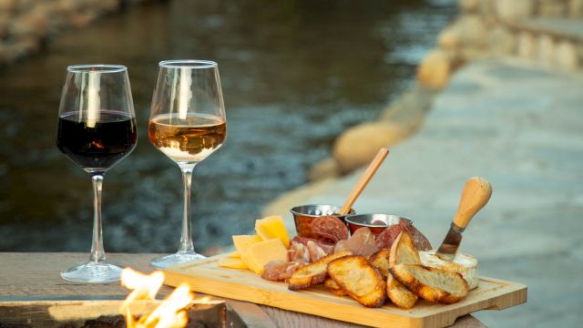 Two glasses of wine sit beside a cheese and meat platter near a fire pit, overlooking a serene stream with stones.