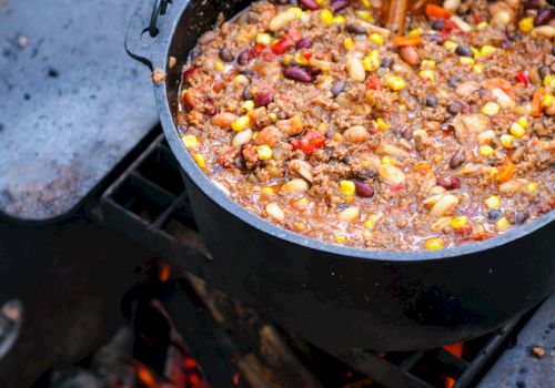 A pot of chili with beans and corn is cooking over an open fire with a wooden spoon in it.