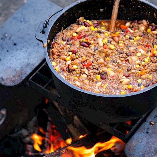 A pot of chili with beans and corn is cooking over an open fire with a wooden spoon in it.