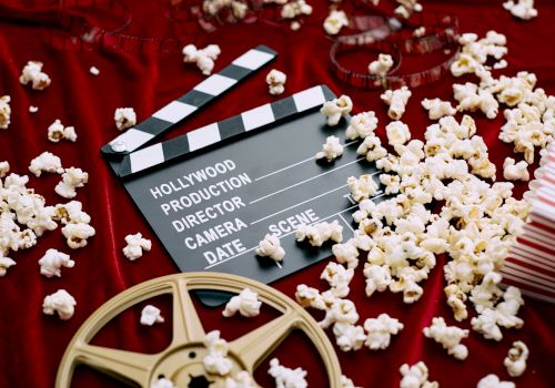 A film reel, popcorn, a clapperboard, and a striped popcorn box on a red fabric surface, suggest a movie theme.