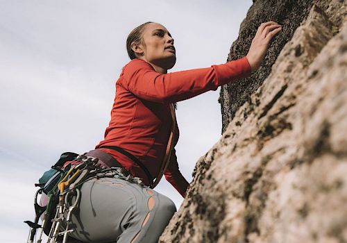 A person in climbing gear is scaling a rock face, focused and determined, with various equipment attached to their harness.