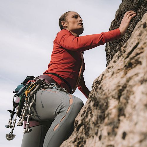 A person in climbing gear is scaling a rock face, focused and determined, with various equipment attached to their harness.