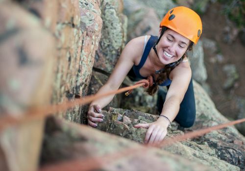 A person wearing an orange helmet is rock climbing, smiling, and holding onto a rope on a rocky surface.