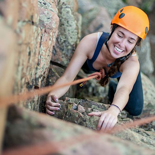 A person wearing an orange helmet is rock climbing, smiling, and holding onto a rope on a rocky surface.