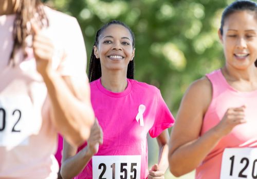 Women are running in a race, wearing numbered bibs and pink shirts with a ribbon symbol, possibly for a breast cancer awareness event.