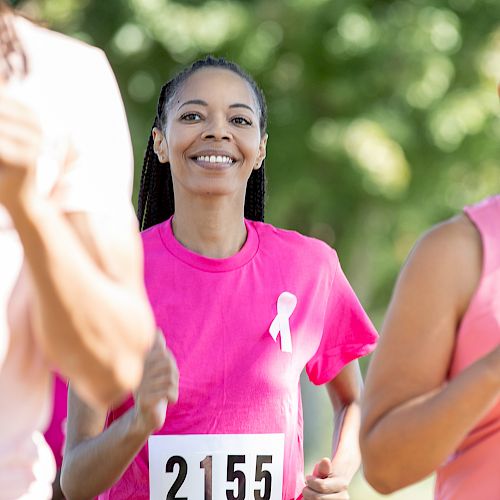 Women are running in a race, wearing numbered bibs and pink shirts with a ribbon symbol, possibly for a breast cancer awareness event.