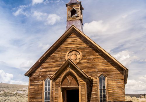 A rustic, wooden church with a steeple stands in a sandy, open landscape under a partly cloudy sky.