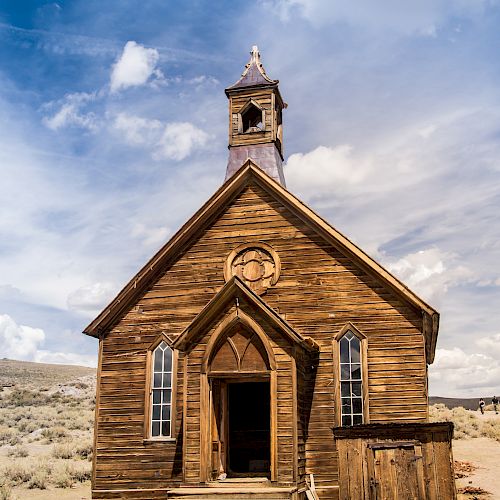 A rustic, wooden church with a steeple stands in a sandy, open landscape under a partly cloudy sky.