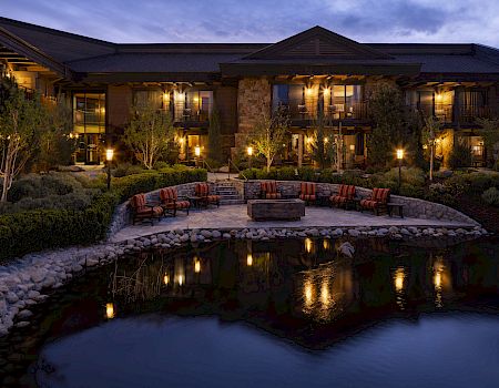 A warmly lit lodge patio with striped chairs around a stone firepit, lush landscaping, and a reflecting pond at dusk.