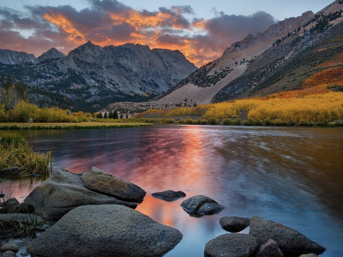 A serene lake with rocks in the foreground, surrounded by autumn foliage, and mountains under a vibrant sunset sky.