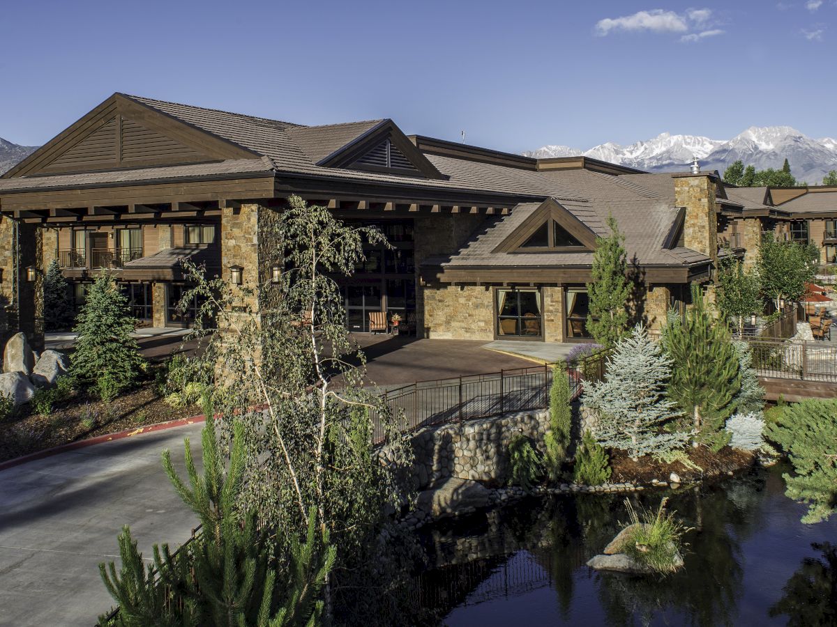 A stone-and-wood lodge with large windows, landscaped gardens, a small pond, and snowcapped mountains in the clear blue sky.