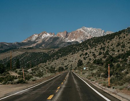 A long empty road winds through green hills toward snowy mountain peaks under a clear blue sky.
