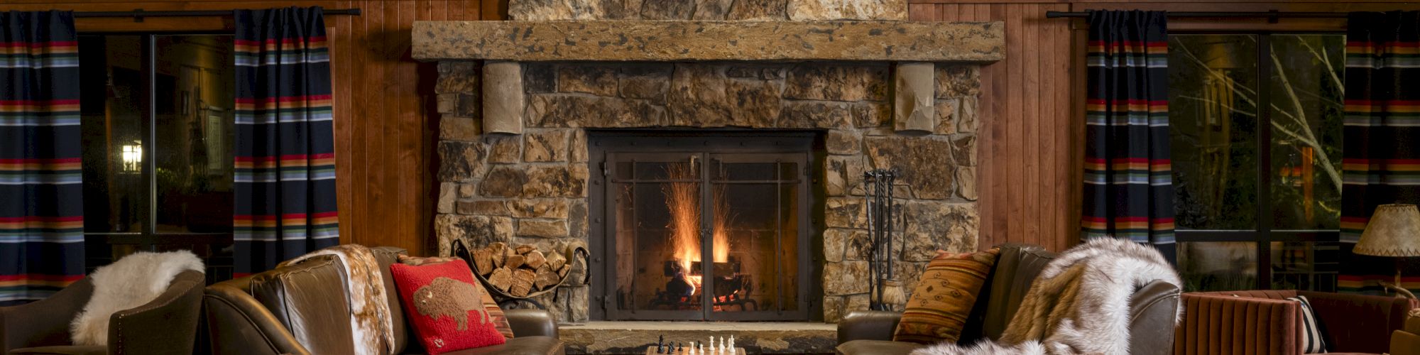 A cozy room with a stone fireplace, surrounded by wooden walls, chairs, a chess board, and warm lighting under a framed picture.
