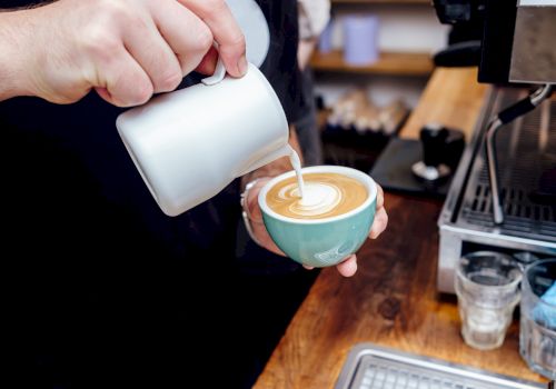 A person is pouring steamed milk into a cup of coffee, creating latte art at a counter alongside an espresso machine.