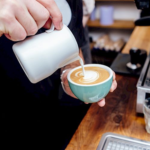 A person is pouring steamed milk into a cup of coffee, creating latte art at a counter alongside an espresso machine.