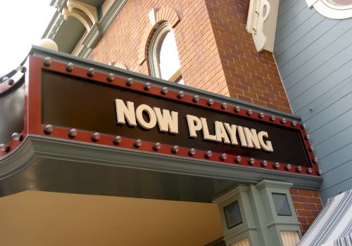 A theater marquee displaying the text "NOW PLAYING" in bold letters, set against a backdrop of brick and painted wood.