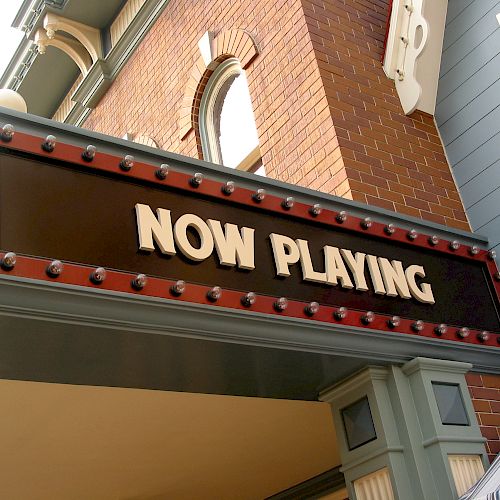 A theater marquee displaying the text "NOW PLAYING" in bold letters, set against a backdrop of brick and painted wood.