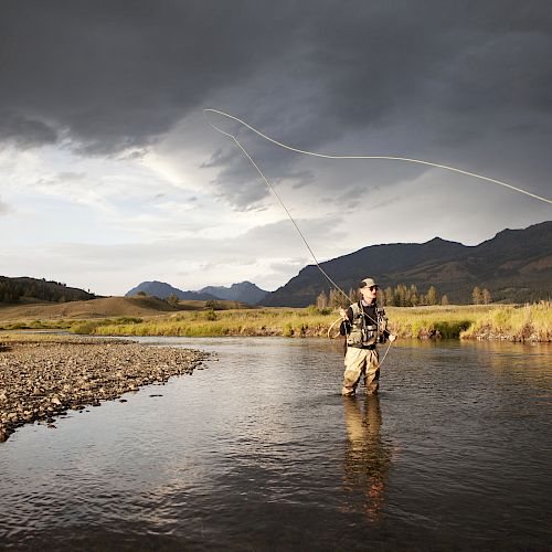 A person is fly fishing in a shallow river under a cloudy sky, surrounded by mountains and grassy fields.